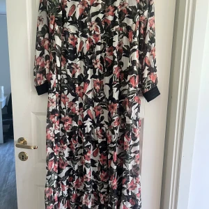 Blommig långklänning från DRYS - En elegant långklänning från DRYS med ett vackert blommönster i rosa, svart och grönt. Klänningen har långa ärmar med svarta muddar vid ärmsluten.