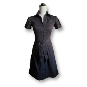 Svart kortärmad klänning med knytband - En elegant svart kortärmad klänning med knytband i midjan. Klänningen har en krage och knappar framtill, vilket ger den en stilren och klassisk look. Fint skick!