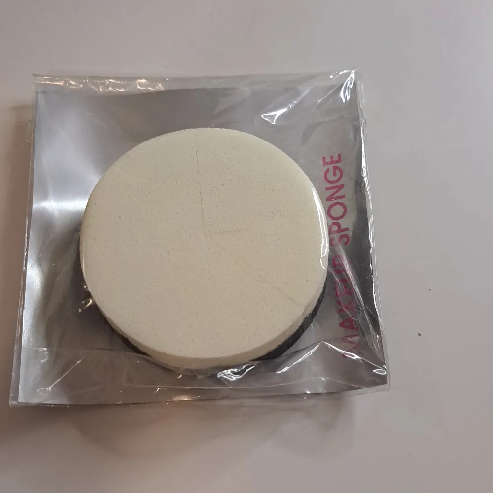 En rund makeup-svamp från Lindex, förpackad i en genomskinlig plastförpackning. Svampen är vit och används för att applicera smink jämnt över ansiktet.. Övrigt.