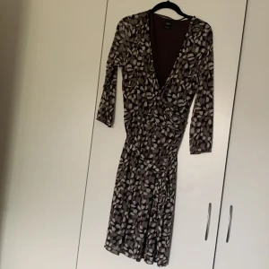 Mönstrad klänning från Esprit - En elegant klänning från Esprit med ett abstrakt mönster i olika nyanser av brunt och beige. Klänningen har en omlott-design med v-ringning och trekvartsärmar. Materialet är lätt och luftigt, vilket gör den perfekt för både vardag och fest.