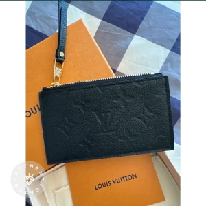 Svart plånbok från Louis Vuitton - Säljer en snygg och elegant svart plånbok från Louis Vuitton. Plånboken är i skinn och har ett diskret mönster med Louis Vuittons ikoniska logga. Den har en dragkedja upptill och en praktisk rem för att enkelt kunna bära den. Perfekt för att hålla dina kort och kontanter säkra med stil!