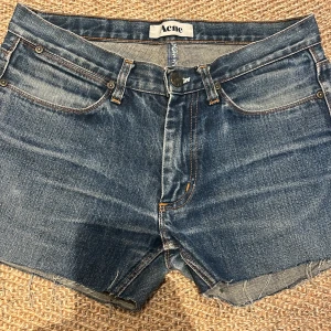 Blå jeansshorts från Acne - Säljer ett par snygga lågmidjade, blå jeansshorts från Acne. De är i bra skick och har en klassisk fyrficksdesign med knapp och dragkedja fram. Shortsen har en rå kant nertill som ger en cool och avslappnad look. Perfekta för sommaren!