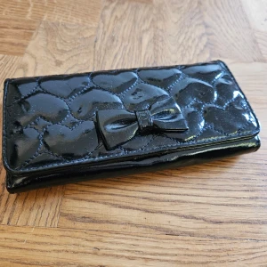 Svart plånbok med rosett - En elegant svart plånbok med glansig yta och quiltat hjärtmönster. Plånboken har en dekorativ rosett på framsidan och flera fack för kort och sedlar inuti. Den stängs med en tryckknapp.