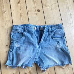Blå jeansshorts med slitningar - Säljer ett par snygga blå jeansshorts från Denim i storlek S. De har en låg midja och är slitna på framsidan för en cool och avslappnad look. Shortsen har fransiga kanter och en knapp- och dragkedjestängning. Perfekta för sommaren!