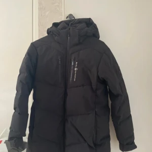 Svart dunjacka från Sail Racing - En svart dunjacka från Sail Racing med huva och dragkedja. Jackan har en logotyp på bröstet och på huvan. Den är designad för att hålla värmen och är tillverkad i Vietnam.