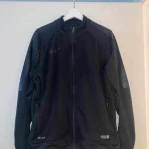 Nike - Svart jacka med dragkedja framtill och två fickor på sidorna. Logotypen är synlig på bröstet och på vänster ärm.