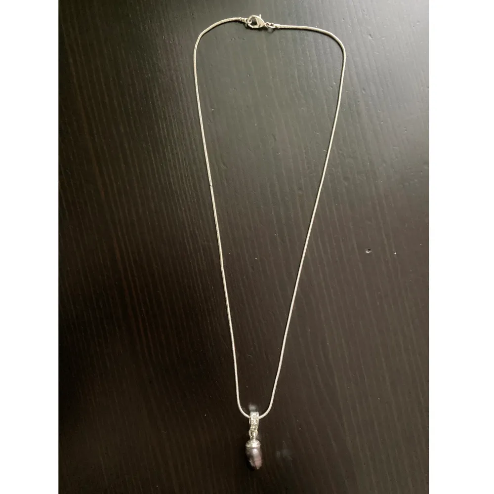 Silverfärgat halsband med lila pärla som hänge samt strassdetaljer. Finns ett hack på pärlan samt på kedjan. Längd: 48,5, bredd: 1 mm, hänge längd: 2,7 cm.. Asusteet.