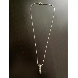 Silverfärgat halsband med lila pärla som hänge samt strassdetaljer. Finns ett hack på pärlan samt på kedjan. Längd: 48,5, bredd: 1 mm, hänge längd: 2,7 cm.