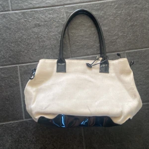 Handväska  - En handväska med svart lack och lite beige strand material eller vad man kan säga med rosetter på svidarna och stor förvaring i. Säljer för 80kr men går att pruta💕💕