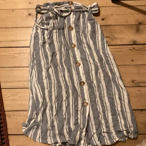 Randig sommarkjol - Medels lång kjol, randig i vitt och blått. Knytband i midjen och stora, träliknande knappar långs mitten av försidan. Storlek 44, men kan passa från 38. 