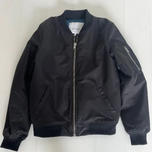 Bomberjacka från Stay, svart, strl S - Svart bomberjacka från märket Stay (inköpt på Carlings) i strl S. Muddar vid halsringning, ärmslut och nedtill. Silverfärgad dragkedja mitt fram samt en liten dragkedja på fickan på ärmen. Petrolblått foder. Mycket fint skick.