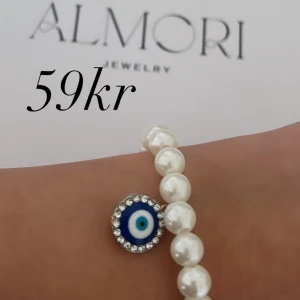 Pärlarmband med öga - Ett elegant pärlarmband från Almori med ett dekorativt öga i mitten. Armbandet består av vita pärlor och har en blå och vit ögondekoration omgiven av små glittrande stenar.