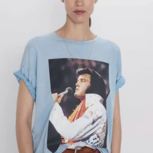 Ljusblå t-shirt med Elvis-tryck från Zara - En ljusblå t-shirt från Zara med ett stort tryck av Elvis Presley på framsidan. Baksidan har texten 'ELVIS' dekorerad med stjärnor och paljetter. T-shirten har korta ärmar och en normal passform.Fint skick, men lite tvättpåverkade och därför blekare i färgen. De har också några små vita fläckar/blekare fläckar. behöver du flera bilder, hör av dig 