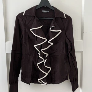 Mörkbrun skjorta Trucco - Skjorta i mörkbrun färg och vitkantiga volanger i storlek 40 från märket Trucco. Skulle säga den är väldigt liten i storleken och gissar snarare på att den passar en S. Är såklart lite skrynklig just nu för den bara legat i garderoben tyvärr.