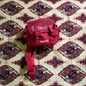 Röd Supreme axelremsväska  - En röd supreme axelremsväska. Helt ny skick. Använd väldigt få gånger. Två remmar så man kan bära på olika sätt (syns på 3 bild). Många fickor. Får plats med massa grejer, går säkert att trycka in en hoodie tex. Mer detaljerade bilder kan skickas 