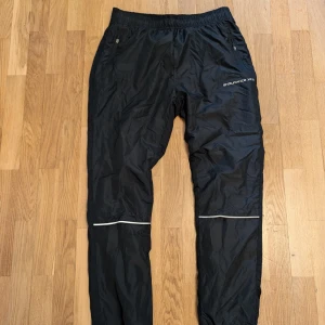 Svarta överdragsbyxor  - Svarta överdragsbyxor med reflexdetaljer. Byxorna har fickor på sidorna och en dragsko i midjan.