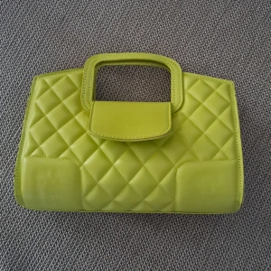 Grön quiltad handväska - En stilren grön handväska med quiltat mönster. Väskan har en rektangulär form och är utrustad med ett handtag. Den har en knappstängning och en liten ficka på framsidan. kommer inte med lång kjedian!! 