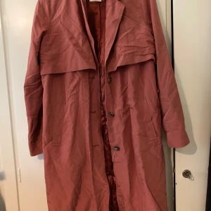 Threnchcoat - Rosa threnchcoat med ett midjeband i samma färg. Den ena hällan för bandet har gått av. 