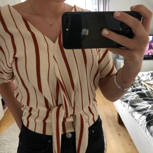 Kort randig blus/tröja med en söt knut - OANVÄND!!! Helt ny blus från Lindex i storlek S. Köpt till ett evenemang som inte blev av. Mycket behagligt tyg. Blusen är beige/orange/brun randig! 