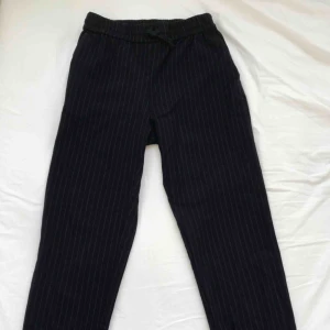  - Kritstrecksrandiga kostymbyxor. Lite kortare modell och inte tighta. Yttre tyget: 65% polyester, 33% viskos, 2% elastan.