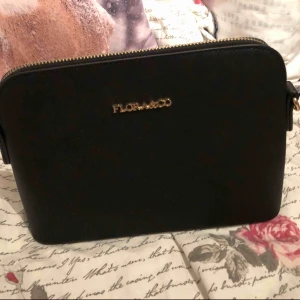  - En jättefin svart väska från flora&co, är i en slags läder imitation med guld detaljer, ett litet fack back en en axelrem medföljer som är justerbart 