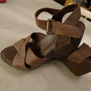  - Ett par sparsamt använda platåsandaler i trä och skinn. Använda tre gånger. 
Storlek 36, med mycket mjuk och vadderad sula. 
Inga skador,  minimalt slitage. 
Skorna mäter 24 centimeter mellan tå och häl. 

Hämtas enligt överenskommelse i centrala Stockholm eller skickas mot frakt i hela Sverige. 
Betalning sker kontant vid träff eller via Swish,  Paypal, Payson.