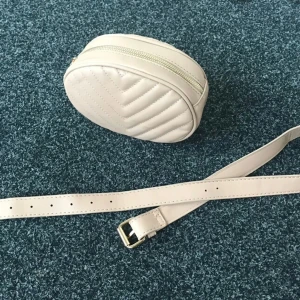 Midjeväska - Vit midjeväska i skinnimitation med guldfärgade detaljer. Väskan har ett svart foder och bältet är ca 100 cm långt.  Väskan är 22 cm bred x 14 cm hög x 6 cm djup. Jag har använt den väska 2 gånger