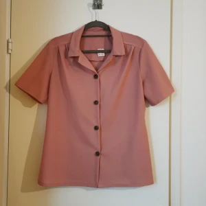 Retro blus i strl 36 - Blus i retrostil från min farmors garderob. Skulle uppskatta den till en liten strl 36, alt en oversize strl 34. Blusen är "gammelrosa" med bruna knappar. 100% polyester. 