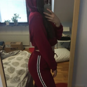  - *OANVÄND*  Tracksuit i två delar. Mörkröd med vit/svarta ränder på benen och luva. Överdelen är ganska kort så som en magtröja och byxorna är medium/high waist. Ganska tunt och väldigt mjukt och bekvämt material!