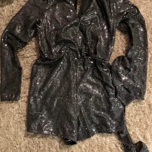 Glittrig playsuit - Glittrig/paljett playsuit storlek 36. Stretchigt material. Har band i midjan att knyta runt. V-ringad.
