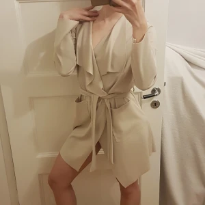 Beige cardigan, stretch material med skärp - Classy beige cardigan trench coat i skönt stretchigt material, med skärp som går att knyta i midjan. Märke: Mirelle. Har några mörka fläckar baktill, bilder kan skickas på dessa på förfrågan. Frakt tillkommer med 48 kr.