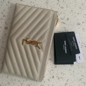 YSL pouch monogram creme/guld i nyskick  - ysl pouch monogram grain de poudre tech i färg créme säljes. Inköpt 2019 för 3800kr. Äkthetsbevis, box, tygpåse och serienummer finns! Nyskick. Dragkedja med 2 fack - ett på framsidan och en ficka invändigt. 