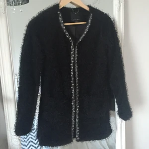  - Svart fluffig/lurvig kofta med fina detaljer av stenar på framsidan längs kanten. Sluts med hyskor framtill. Två rymliga fickor framtill. Mycket bra skick! 