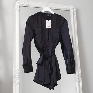 Svart glansig tunika BikBok XS - Färg: svart.  Strl: XS.  Blus/tunika från Bikbok. Helt ny, med lappar kvar. Tillhörande band medföljer. Ger snygg form! Sluts igen med knappar framtill. Halv-lång modell, som går över rumpan. Snygg till både fest & vardags!