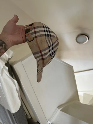 Beige Burberry keps med rutmönster - Säljer en klassisk beige keps från Burberry med det ikoniska rutmönstret i svart, vitt och rött. Justerbar rem baktill i samma tyg. Perfekt accessoar för dig som vill ha en tidlös och trendig look.
