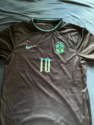 Brasilien fotbollströja Nike nr 10 - Snygg mörkbrun Brasilien fotbollströja från Nike med ljusgröna detaljer, nummer 10 på bröstet och CBF-emblem. Tillverkad i Dri-FIT material som andas och håller dig torr under matchen. Rund hals med ljusgrön rand och korta ärmar.