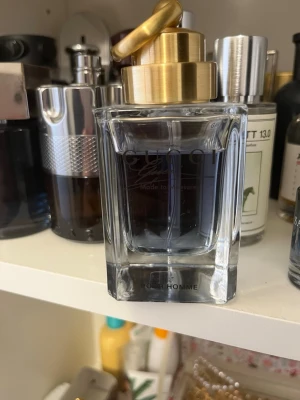 Gucci Pour Homme parfym - Gucci Pour Homme parfym i en elegant, fyrkantig glasflaska med guldfärgad metallkork. Flaskan har en stilren design och innehåller en blåaktig vätska. Perfekt för dig som gillar exklusiva och klassiska dofter.