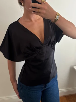 Svart omlottopp i satin-look - Stilren svart omlottopp med djup v-ringning och korta, vida ärmar. Toppen har en elegant satin-liknande finish som ger en lyxig känsla. Den hade band vid midjan som jag klippt bort men det märks inget 