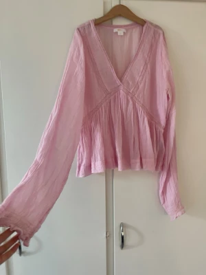 Ljusrosa v-ringad blus - Söt ljusrosa blus med v-ringning och avslappnad passform. Blusen har långa ärmar och är tillverkad i ett tunt, luftigt tyg som ger en bohemisk känsla. Perfekt att styla med jeans eller kjol för en trendig look.