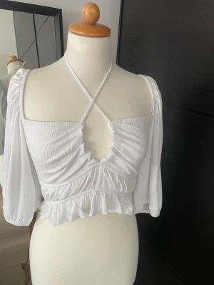 Vit croppad blus med volang från Cubus  - ALDRIG ANVÄND!! Superfin vit croppad blus med långa puffärmar, volangkant och snygg omlott-detalj framtill. Toppen har knyt runt nacken och en smockad rygg för extra komfort.