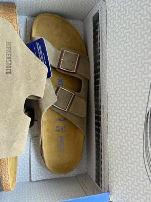Birkenstock Arizona sandaler beige mocka - Säljer ett par Birkenstock Arizona sandaler i beige mocka med två breda remmar och justerbara metallspännen. Klassisk korksula och mjuk fotbädd för extra komfort. Perfekta för dig som vill ha både stil och bekvämlighet i vardagen.