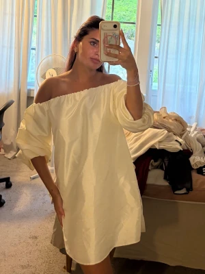 Offshoulder ljus kort klänning - Trendig offshoulder klänning i ljus, nästan vit ton. Klänningen har en lös passform, ballongärmar och är tillverkad i ett glansigt material som påminner om satin. Perfekt för dig som vill ha en enkel men snygg look.