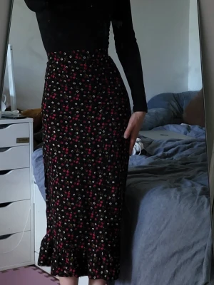 Svart blommig vintage långkjol med volang - Säljer en svart långkjol med små blommor i rött, vitt och rosa. Kjolen har hög midja och en lätt utsvängd form med volang längst ner. Perfekt för dig som gillar romantiska och feminina detaljer.
