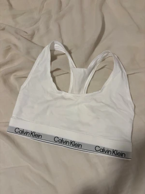 Vit bh från Calvin Klein - Säljer en vit från Calvin Klein med bred resår och logga runt om. Bh:n har brottarrygg och är tillverkad i mjukt, stretchigt material som ger en bekväm passform. Perfekt för dig som gillar enkel och stilren design.