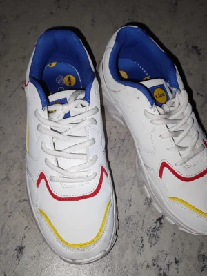 Lidl sneakers i vitt med färgglada detaljer - Säljer ett par vita Lidl sneakers med blå, röda och gula detaljer. Skorna har en chunky sula, blå insida och Lidl-logga på hälen och innersulan. Tillverkade i syntetmaterial och textil. Perfekta för dig som gillar färg och vill sticka ut.
