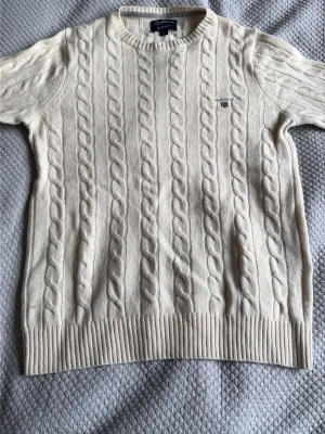 Vit kabelstickad tröja från Gant - Kabelstickad tröja i offwhite från Gant med rund halsringning och ribbade muddar. Tröjan har ett diskret Gant-broderi på bröstet och är tillverkad i mjukt material. Perfekt för en klassisk och stilren look.