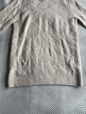 Beige stickad tröja med strukturmönster - Säljer en beige stickad tröja med snyggt strukturmönster och rund hals. Tröjan har långa ärmar och ribbade muddar vid ärmslut och nederkant. Perfekt för dig som gillar stilrena och mysiga plagg i neutrala färger.