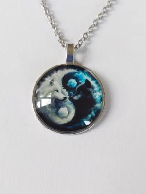 Halsband med yin yang vargar - Unikt halsband med runt hängsmycke i metall. Motivet visar två vargar, en vit och en svart, i yin yang-stil mot en blå bakgrund, I 2D.Kedjan är silverfärgad och länkarna är tunna. Materialet rostfritt stål 