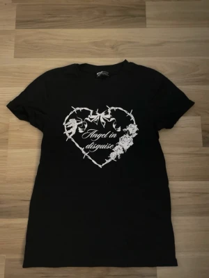 Svart t-shirt med hjärtmotiv - Svart t-shirt från Bershka med vitt tryck av ett hjärta av taggtråd, rosor och texten 'Angel in disguise' på bröstet. Klassisk passform och rund halsringning. Perfekt för dig som gillar edgy och stilrena plagg.