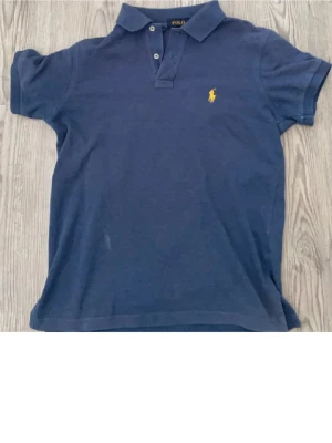 Blå pikétröja från Polo Ralph Lauren - Klassisk blå pikétröja från Polo Ralph Lauren med gul broderad logga på bröstet. Tröjan har korta ärmar, krage och knappar vid halsen. Tillverkad i bomull och har en normal passform. Perfekt till en avslappnad men ändå stilren look.